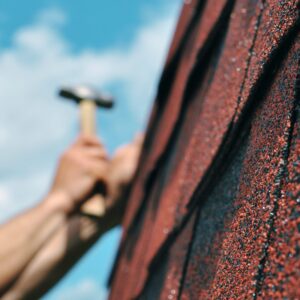 Close-up of red asphalt shingles with a roofing professional in the background securing the roof with a hammer. Ryan Construction Systems provides expert roofing services and shingle replacement in Kansas City, Parkville, Liberty, Gladstone, and nearby areas. Your trusted local roofing contractor for residential roof repair and installation near you.