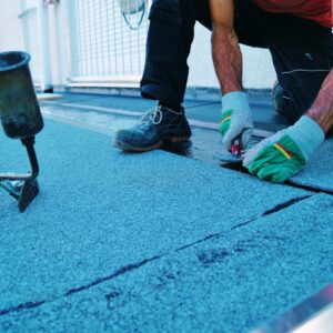 Roofer installing waterproof membrane on a flat roof surface using adhesive and hand tools during residential roof construction.
