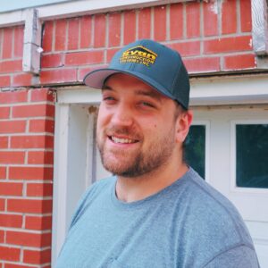 A smiling roofing contractor wearing a Ryan Construction Systems hat, standing in front of a brick home.
